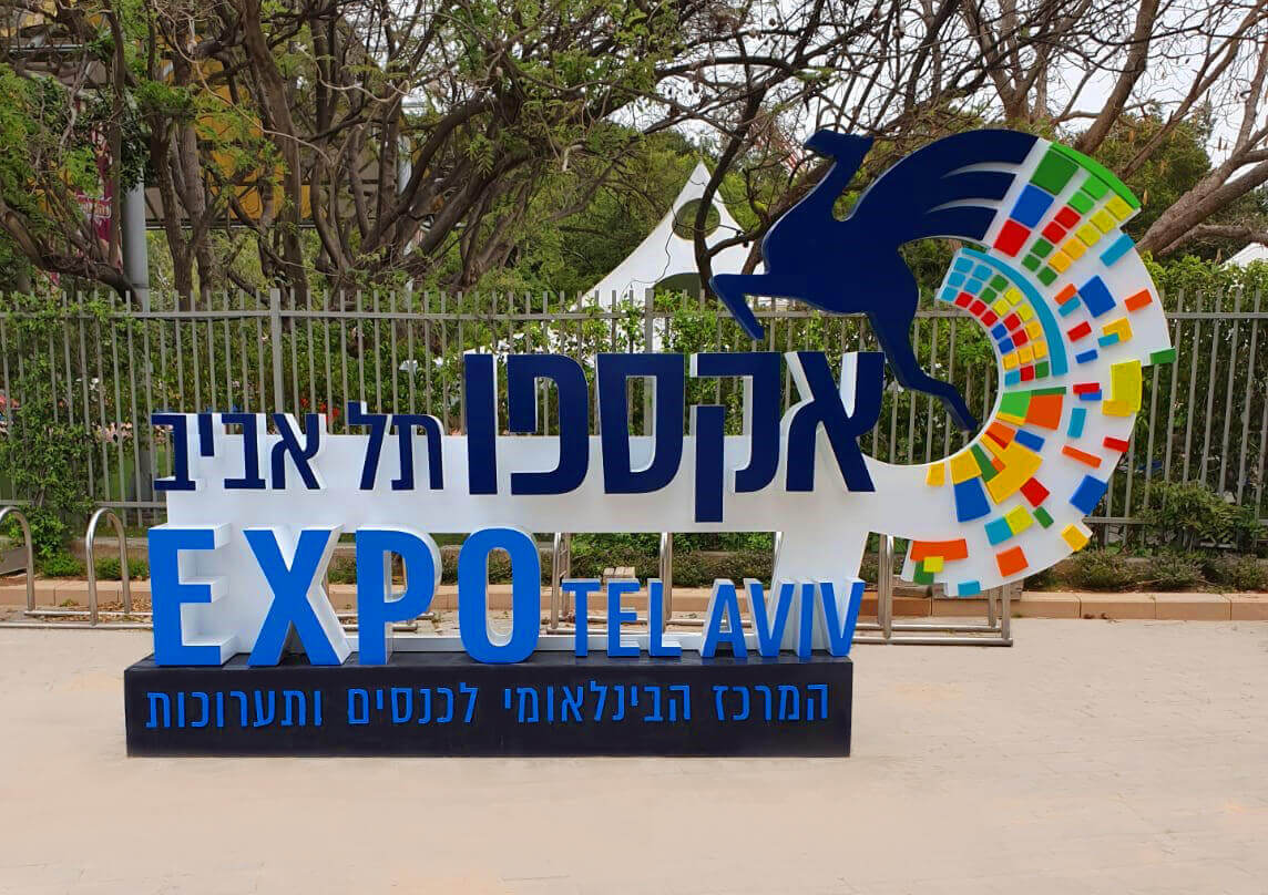 צילום של שלט המיתוג החדש "אקספו תל אביב EXPO TEL AVIV" הכולל את הלוגו המעודכן עם סמל הגמל המעופף בגרסה מודרנית וצבעונית. השלט מוצב בכניסה למתחם ומסמל את הפיכתו למרכז כנסים ותערוכות בינלאומי.